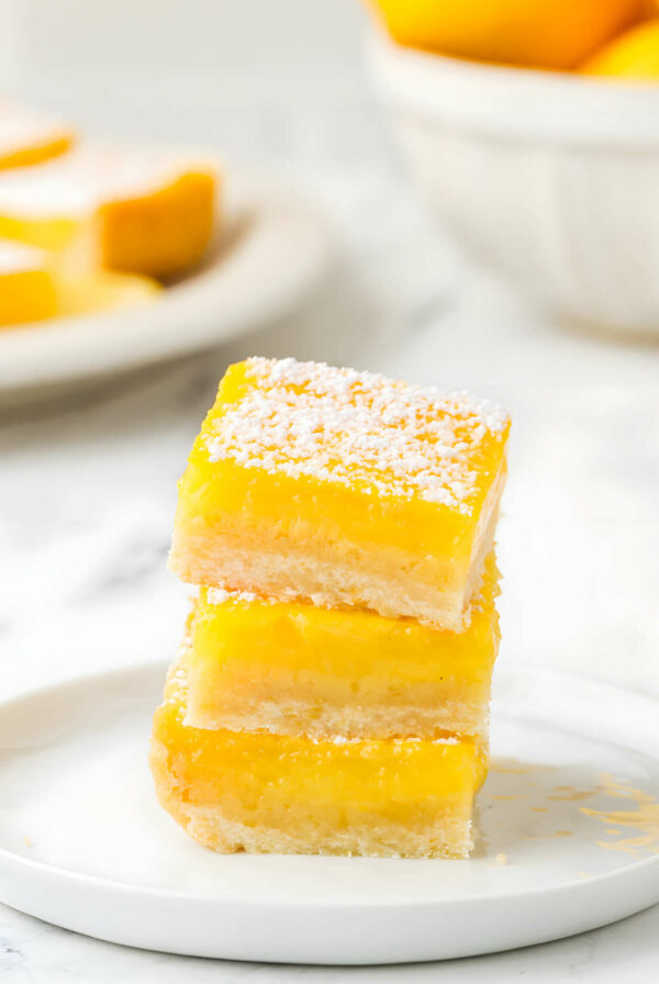 Three lemon bars stacked on a white plate with a bowl of lemons in the background.