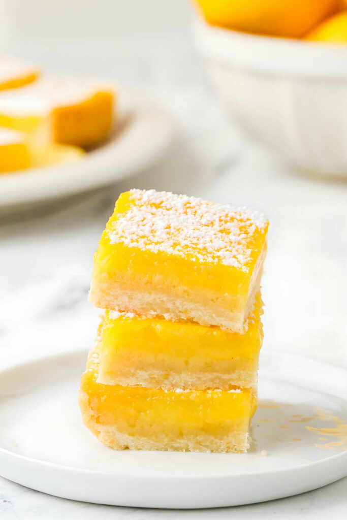 Three lemon bars stacked on a white plate with a bowl of lemons in the background.
