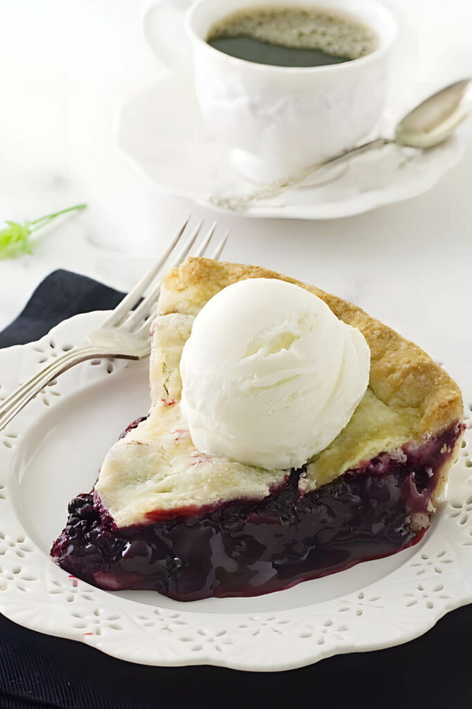 Blackberry pie with vanilla ice cream on a plate, coffee cup in background.