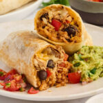 A chicken burrito cut in half and on a plate with guacamole.