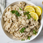 Cooked shredded chicken in a bowl with green onions and lemon slices for garnish.
