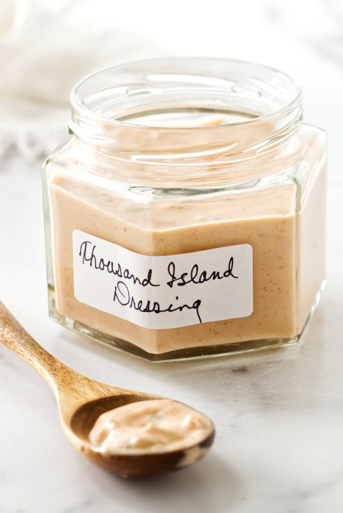 Jar of Thousand Island dressing on marble, wooden spoon with dressing in front.