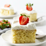 Frosted three milk cake slice with strawberry on white plate and fork.