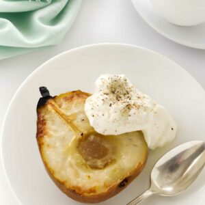 Half a baked pear with cream and pepper on a white plate beside a spoon.