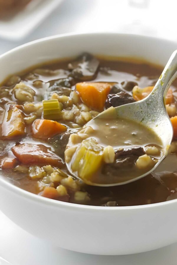 Spoon lifting beef and barley soup with vegetables in a bowl.