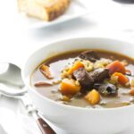 Bowl of beef and vegetable soup with bread and red wine in background.