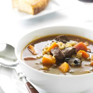 Bowl of beef and vegetable soup with bread and red wine in background.