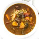 White bowl of beef and vegetable soup with barley, on plate with spoon.