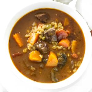 White bowl of beef and vegetable soup with barley, on plate with spoon.