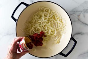 Hand pours tomato paste into white pot with onions on marble countertop.