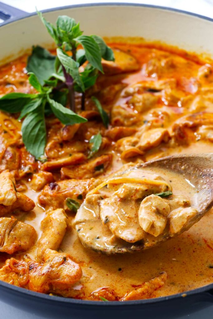 Close-up of creamy panang curry chicken in a pan, basil garnish, wooden spoon.