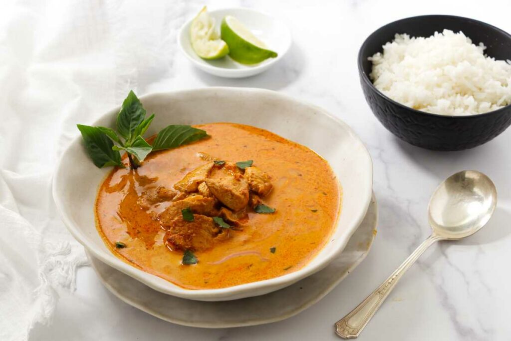 Panang curry with chicken, basil garnish, rice, and lime wedges on white surface.