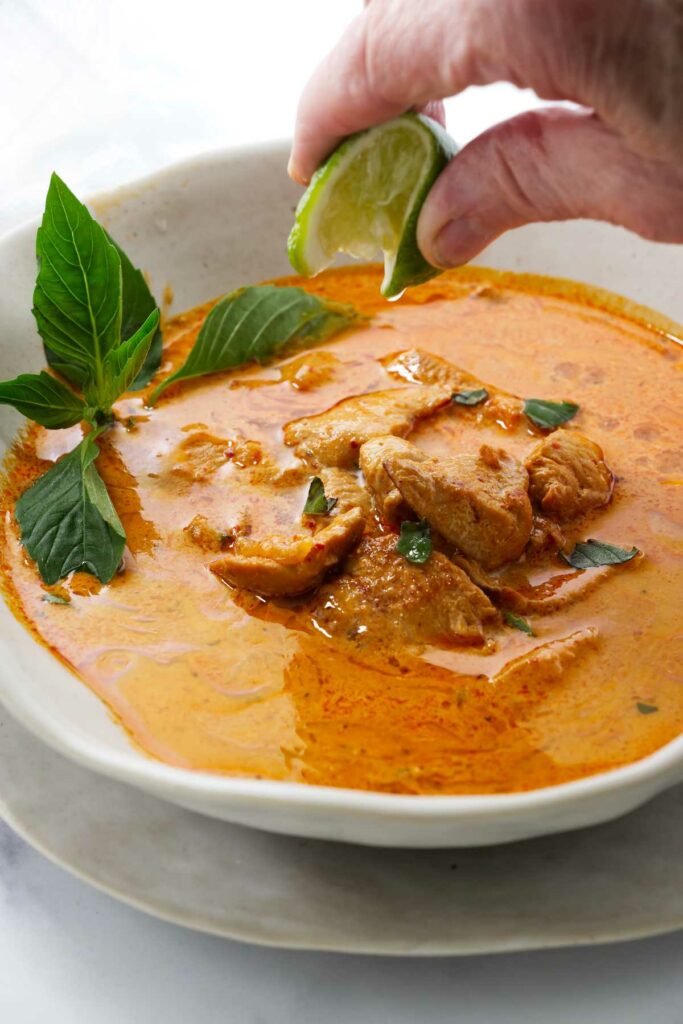 Hand squeezing lime over chicken curry, garnished with basil leaves.