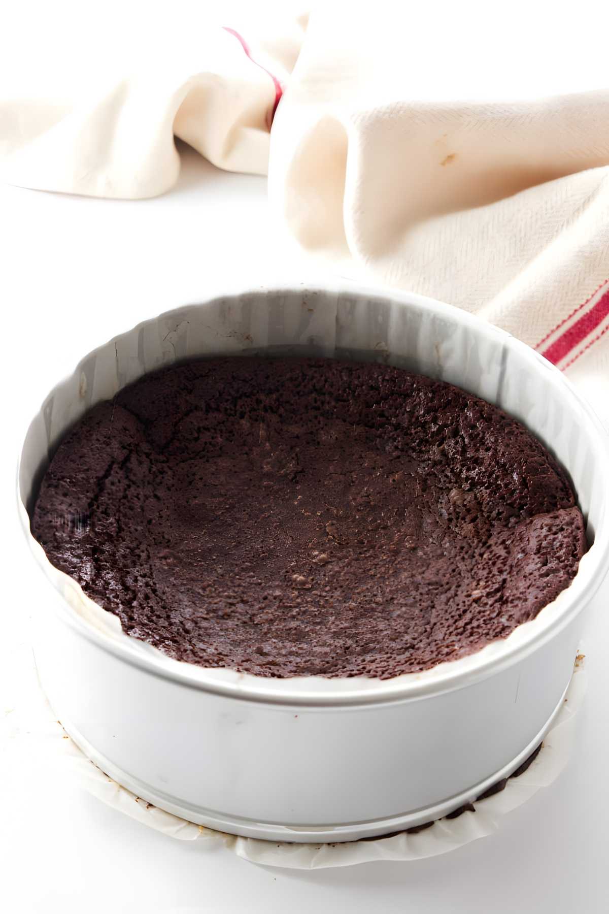 Round dark chocolate cake in springform pan on white surface with striped cloth.