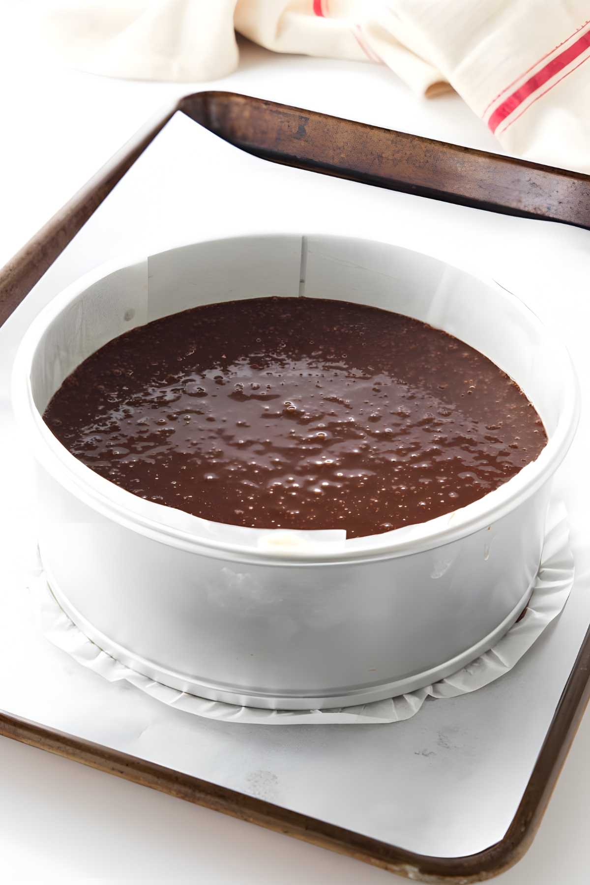 Round pan with chocolate cake batter on baking sheet, light cloth behind.