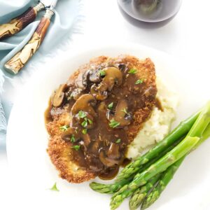 Breaded chicken with mushroom gravy, mashed potatoes, asparagus, and red wine on table.