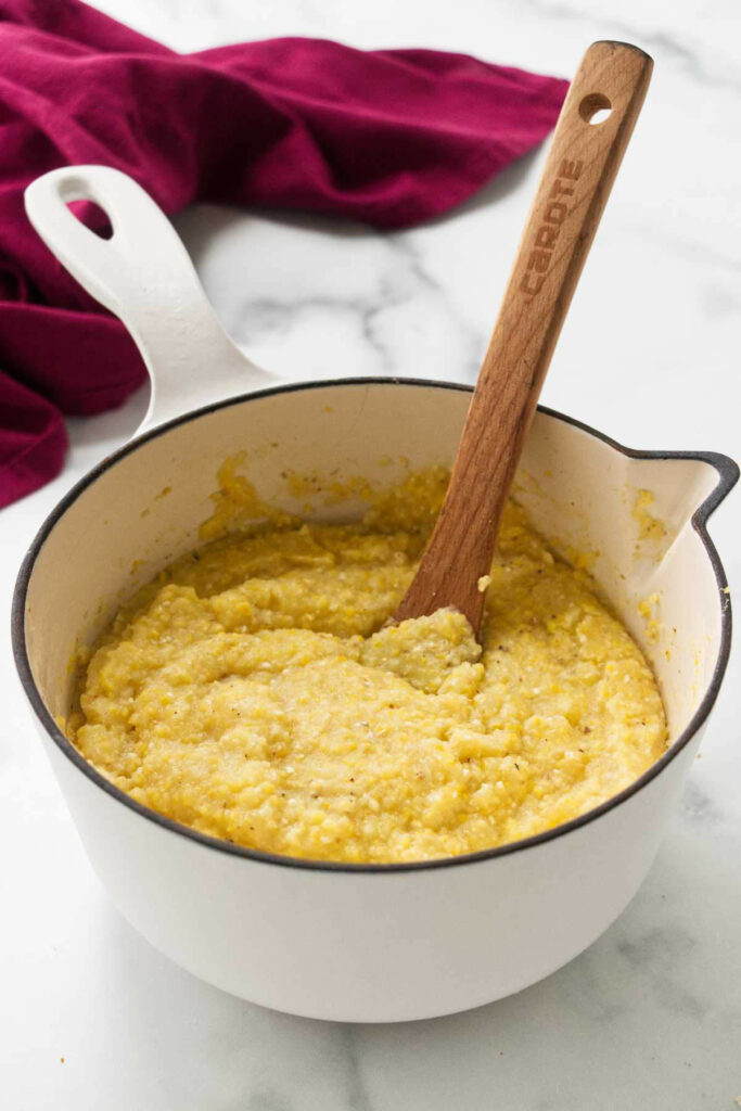 Pot with yellow polenta and wooden spoon on marble counter.