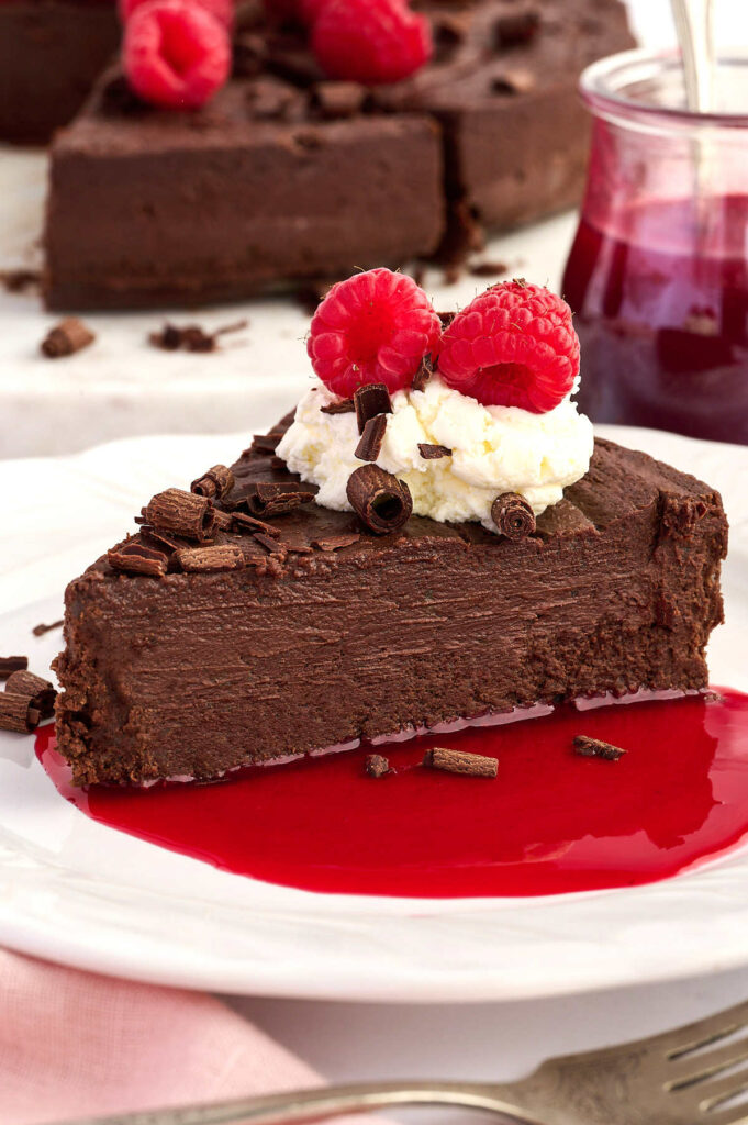 Chocolate torte slice with whipped cream, raspberries, and sauce on a white plate.