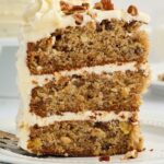Three-layer cake slice with white frosting and nuts on a plate with fork.