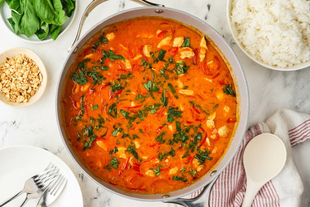 Pan of orange-red curry with herbs, rice, peanuts, and basil on marble.