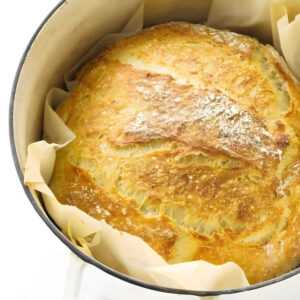 Round, golden-brown crusty bread in a white Dutch oven on parchment.
