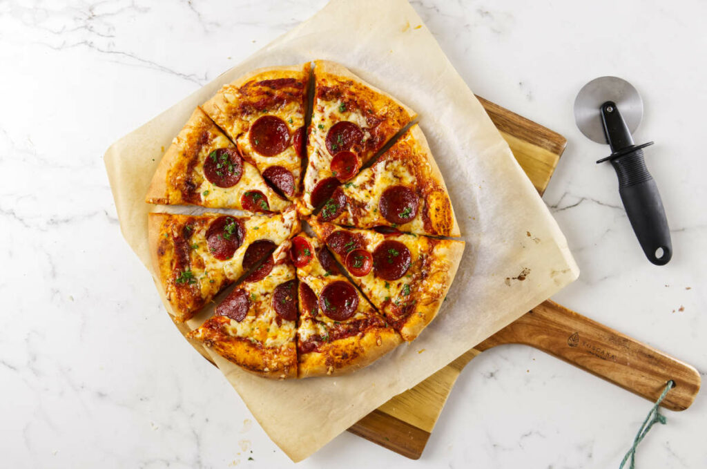 Sliced pepperoni pizza on wooden peel beside a pizza cutter.