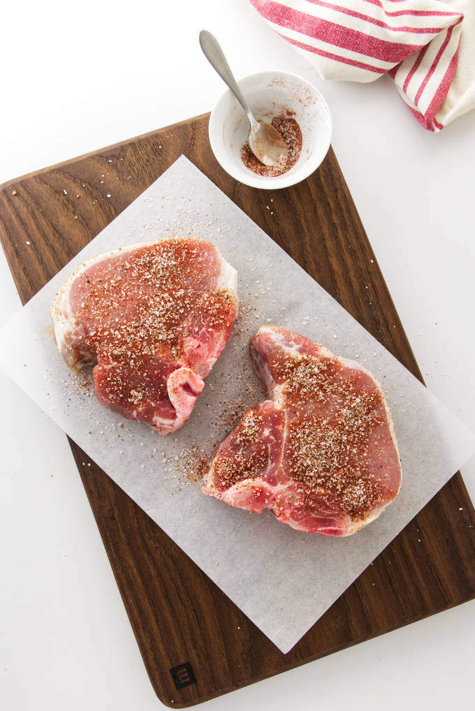 Adding seasoning to two thick pork chops on a cutting board.