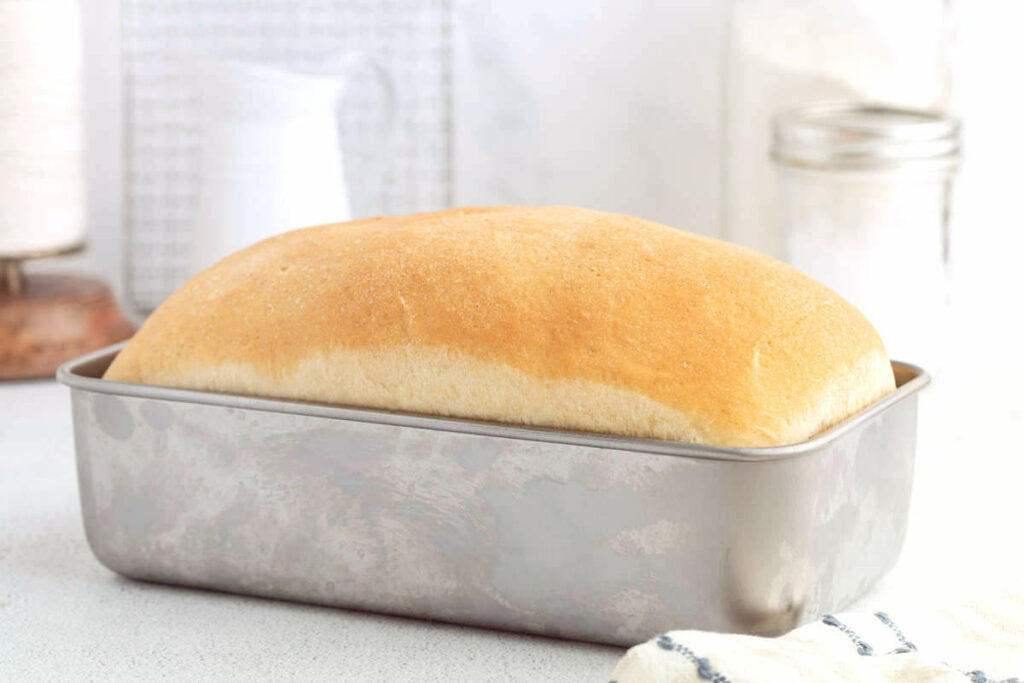 A golden-brown loaf of bread in a metal pan on a white surface.