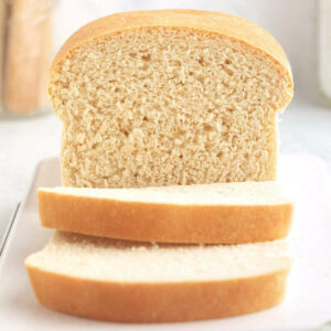Homemade sandwich bread with golden crust and two slices on white surface.