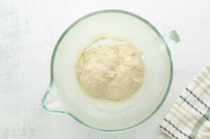Glass bowl with dough on light surface, beside a striped kitchen towel.