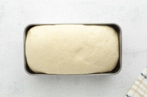 Risen bread dough in loaf pan on light surface, striped towel in corner.