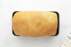 Golden-brown loaf of bread in a baking pan on white surface, cloth at corner.
