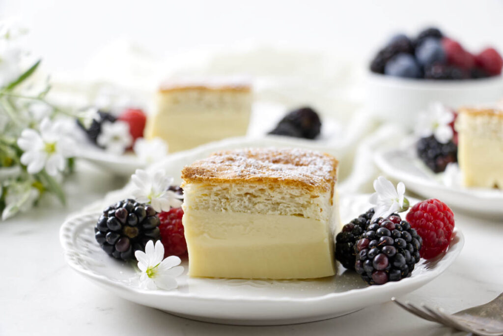 A slice of magic custard cake on a white plate with berries and small white flowers.