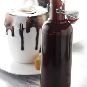 Glass bottle of chocolate syrup with hot chocolate and whipped cream behind.