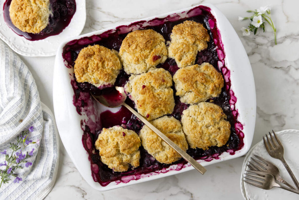 Blueberry cobbler recipe with spoon in the baking dish and a serving plate nearby.