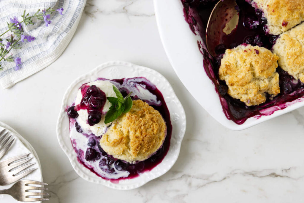 Plated blueberry cobbler with vanilla ice cream beside the baking dish.