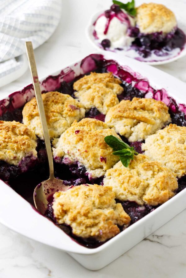 Spoon lifting warm blueberry cobbler from a baking dish with biscuit topping.