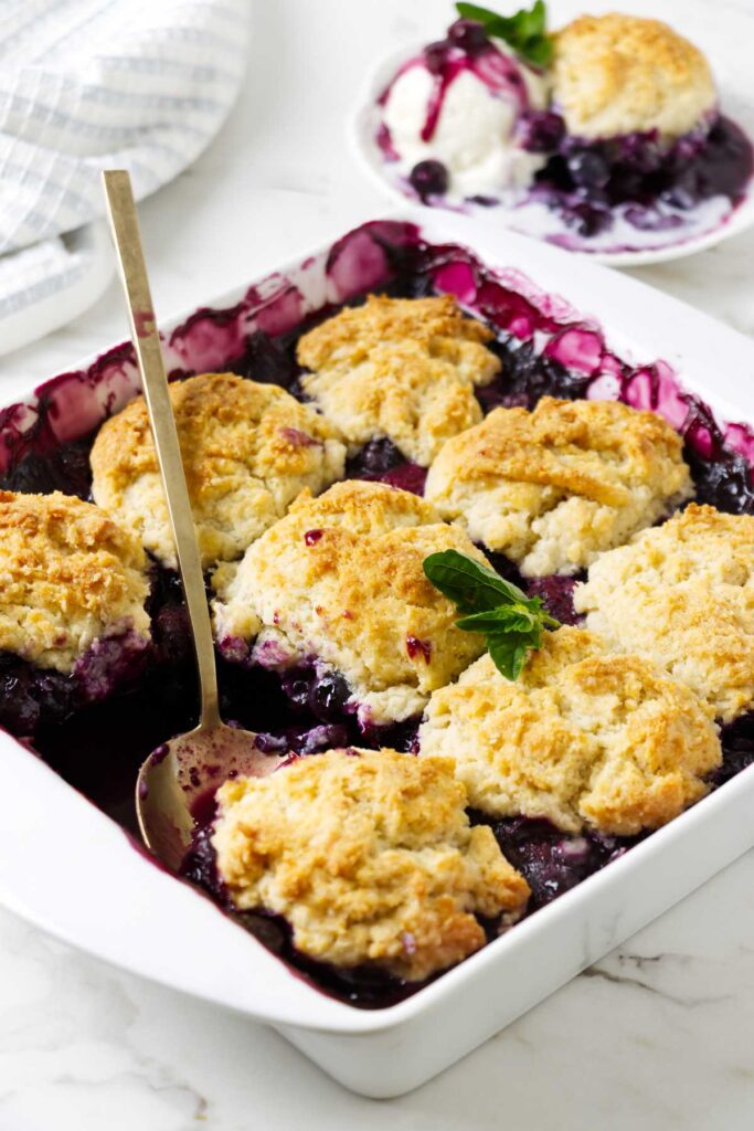 Spoon lifting warm blueberry cobbler from a baking dish with biscuit topping.