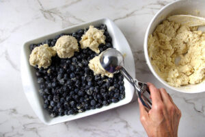 Scooping biscuit dough over blueberry filling for a cobbler recipe.