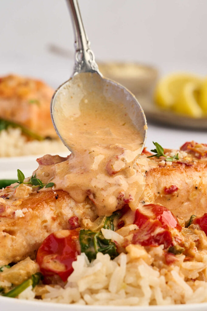 Spoon pouring creamy sauce over skillet salmon with rice and greens.