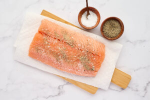 A salmon fillet on a cutting board sprinkled with salt and pepper.