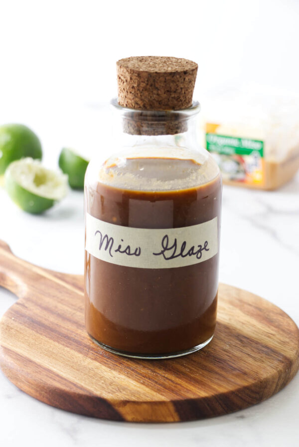 Glass bottle labeled Miso Glaze on wooden board with blurred limes and cheese.