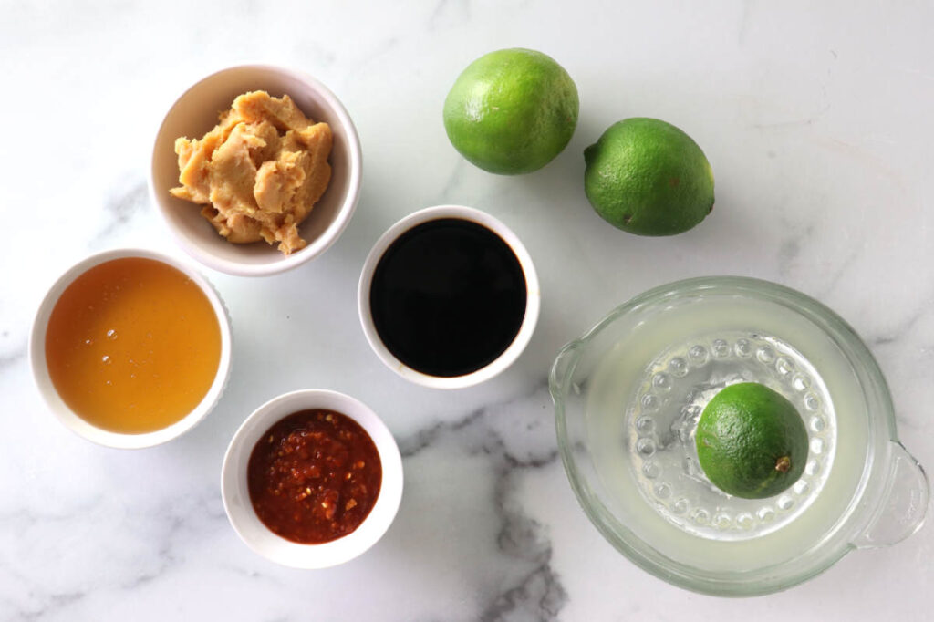 Five small bowls with condiments and limes on a white marble surface.
