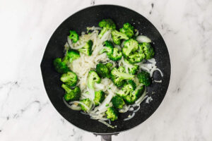 Broccoli florets and sliced onions cooking in a skillet for stir fry.