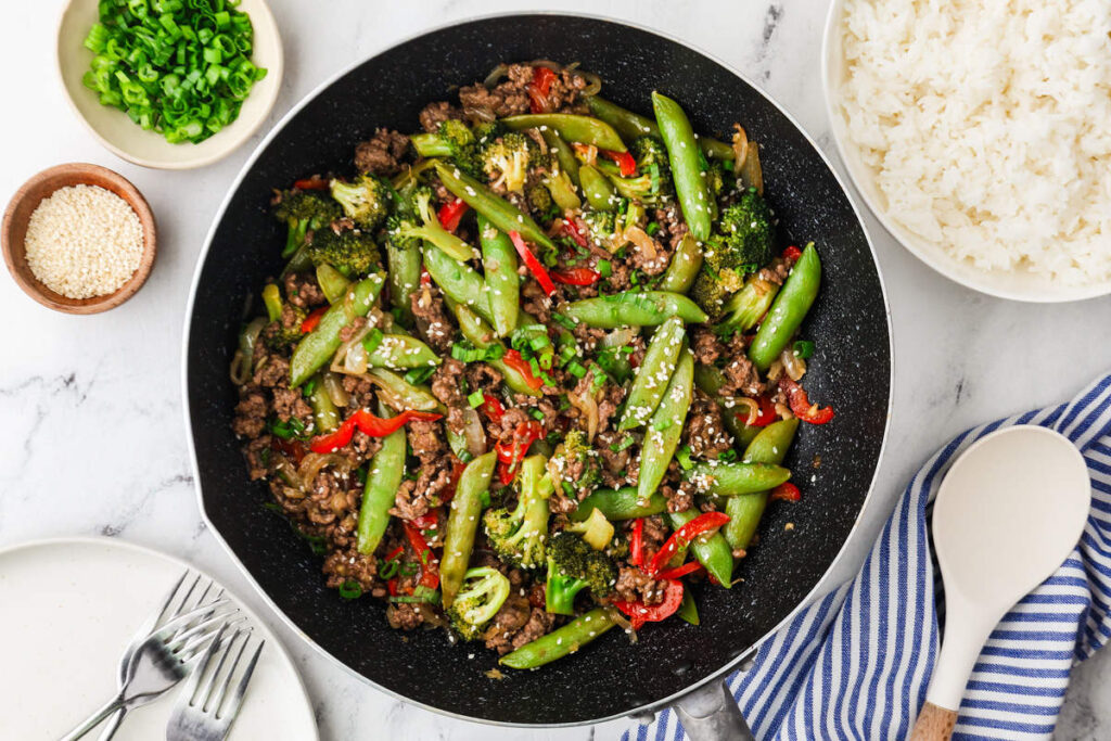 Ground beef stir fry in a skillet with rice and garnishes on the side.