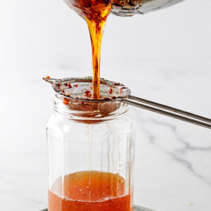 Warm honey pours through strainer with chili flakes into a glass jar.