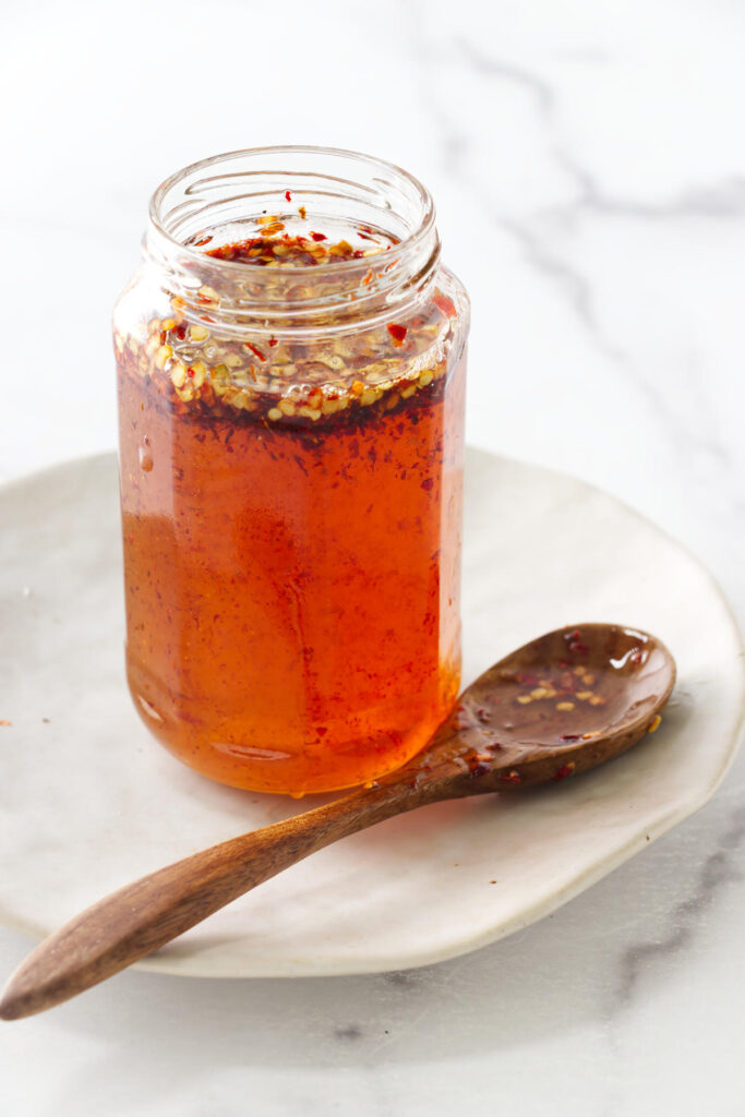 Spicy honey with dried pepper flakes in a jar next to a wooden spoon.