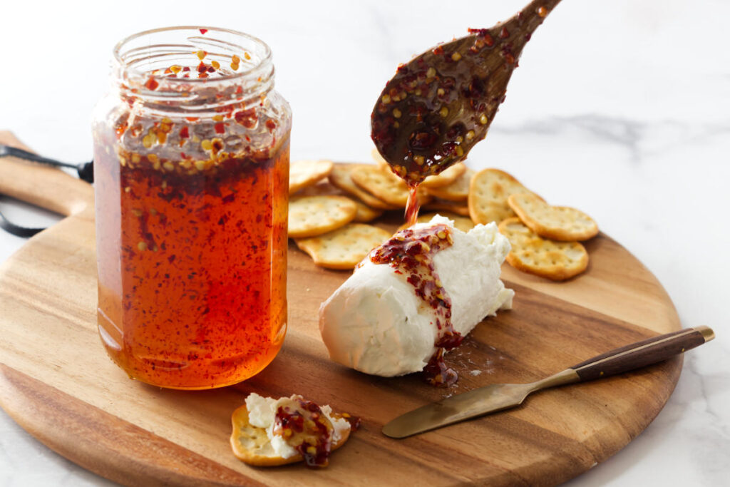 Goat cheese log with hot honey, crackers, and utensils on wooden board.