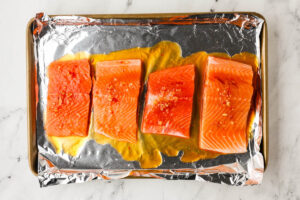 Four raw salmon fillets with sticky sauce on foil-lined baking sheet.