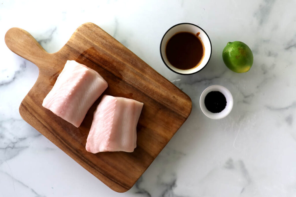 Two raw fish fillets on a wooden cutting board with sauce, pepper, and lime.